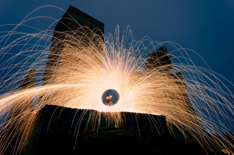 In this photograph titled "First Sparks", I captured an unplanned long exposure experiment from my early days. After work, a friend and I had nothing to do, so we decided to try something new and drove to a nearby castle ruin. In the center, my friend is spinning burning steel wool, creating a glowing circular portal that sprays brilliant orange sparks in dramatic arcs, contrasting with the dark silhouette of the ancient fortress and the deep blue twilight sky.