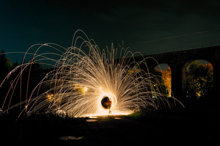 A long-exposure night photograph titled Imprint No.012.jpg, capturing a burning steel wool performance. In the center, a bright circle of light propels hundreds of golden sparks in high, symmetrical arcs against a dark night sky. In the background, a massive stone viaduct with illuminated arches stands silently, framed by dark trees and a wet ground reflecting the fire.