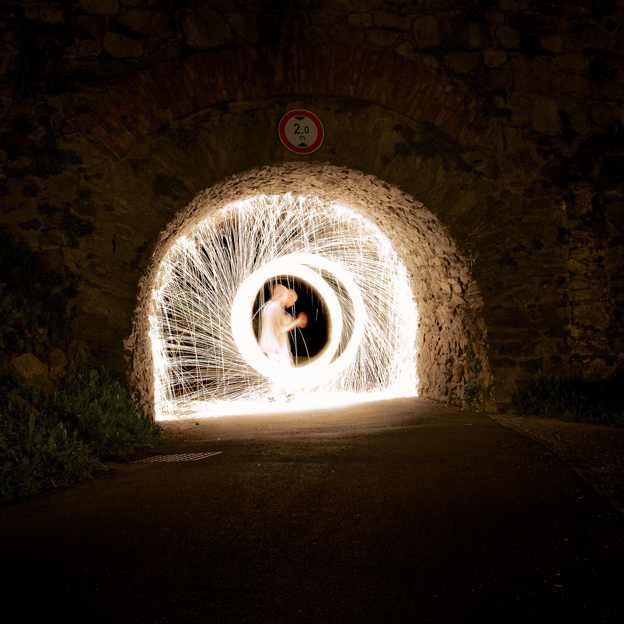 A long-exposure night photograph titled Imprint No.017.jpg. Inside a stone underpass, a figure is spinning burning steel wool, creating a perfect circular tunnel of light. Brilliant orange sparks bounce off the walls, contrasting sharply with the night's darkness and the rough texture of the stone.