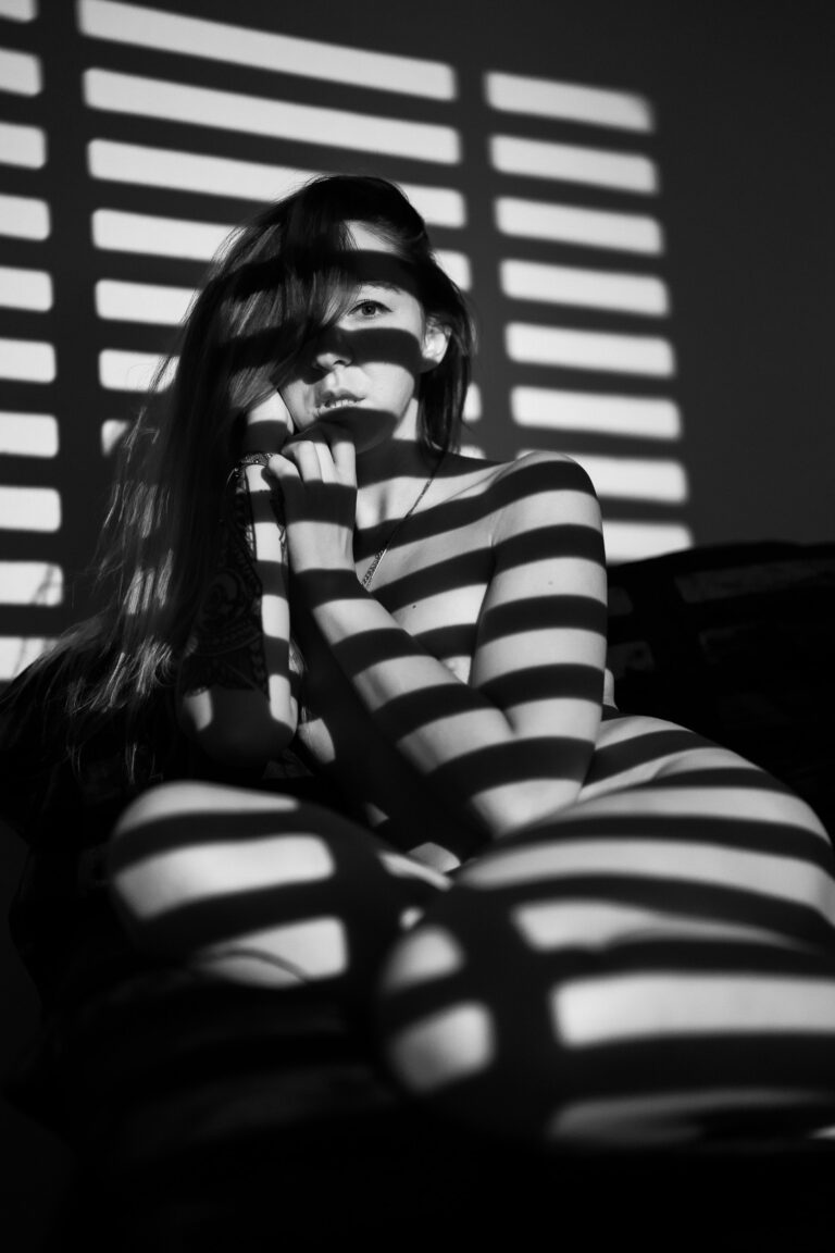 Black and white fine art portrait of a woman, Zoe, sitting in the shadows while geometric light patterns from window blinds project across her body and face.