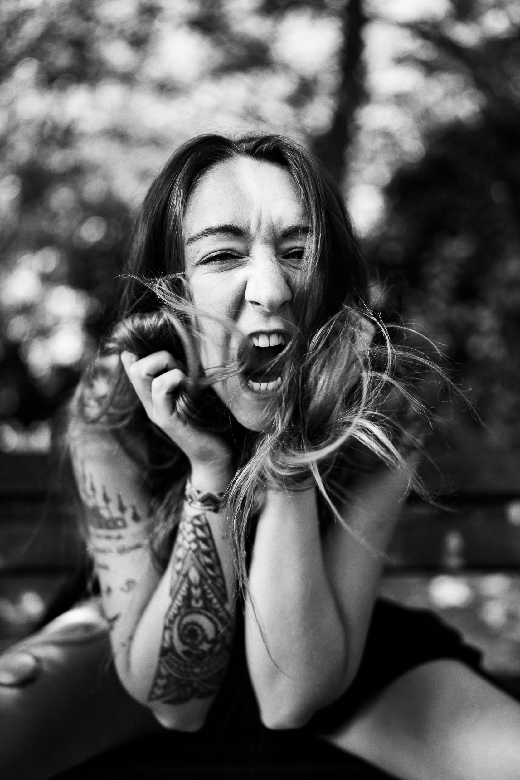 High-contrast black and white close-up portrait of a tattooed woman (Zoe) holding her hair around her face, screaming with raw emotion and energy on a park bench. Background features soft bokeh from trees.