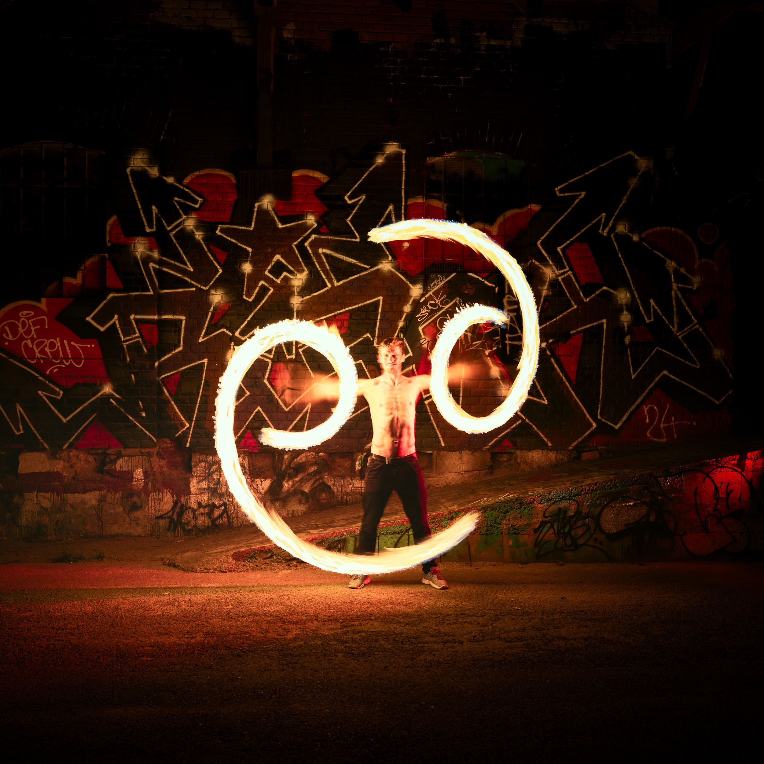 A long-exposure night photograph of a shirtless young man spinning a fire show (poi) on an empty street in front of a graffiti-covered wall. The fire trails create brightly glowing fiery spirals forming the number 96 on each side. The background is dramatically illuminated by the red and yellow light of the fire.