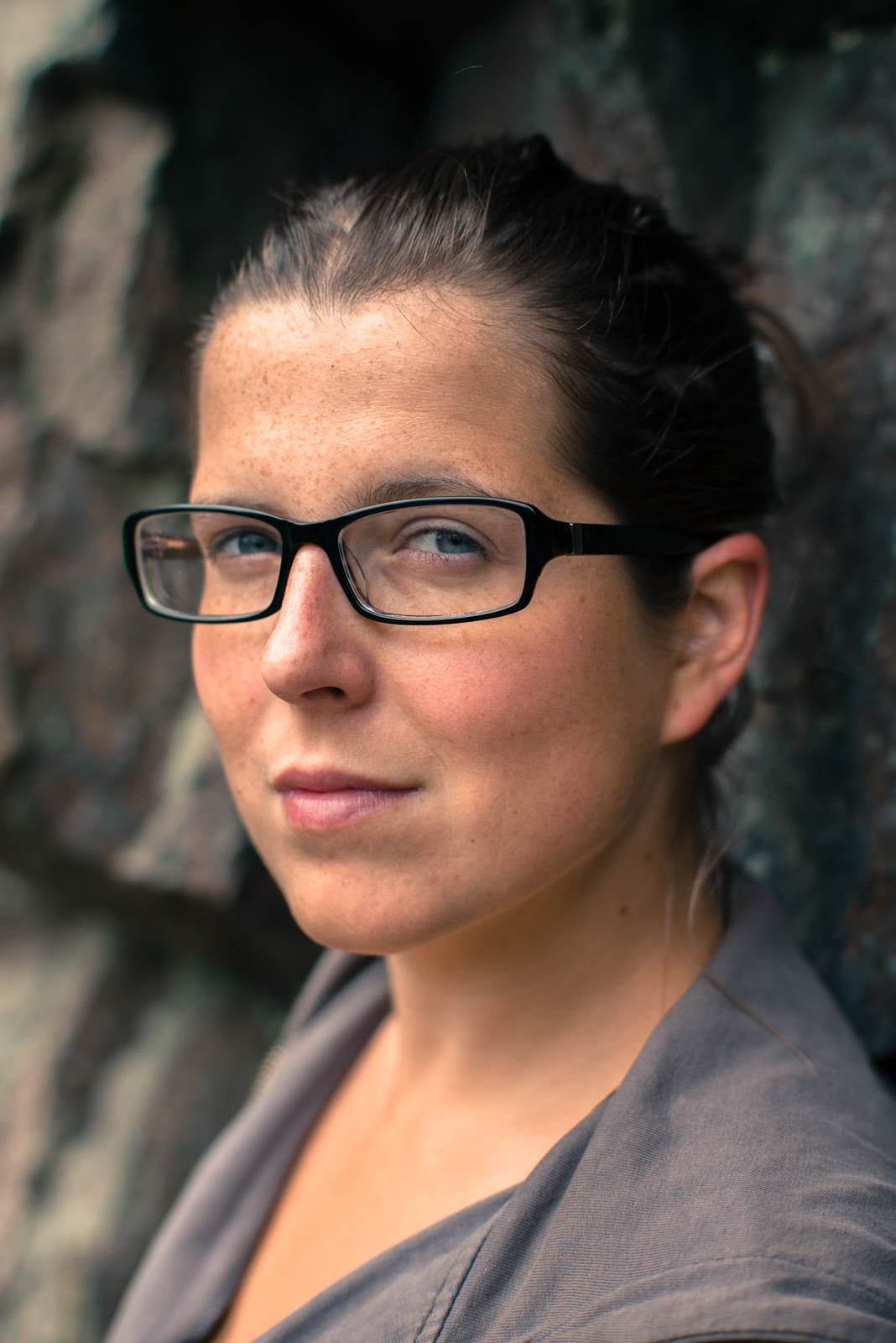 Close up portrait of a woman wearing glasses with freckles on her face, leaning against a stone wall in natural light.